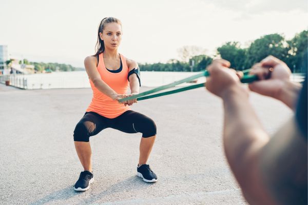 Eine Frau macht eine Kräftigungsübung mit einem Trainingsband, zwei Hände halten das Band gegenüber fest.